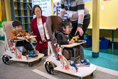 two young children riding their mobility vehicles with aides looking ong